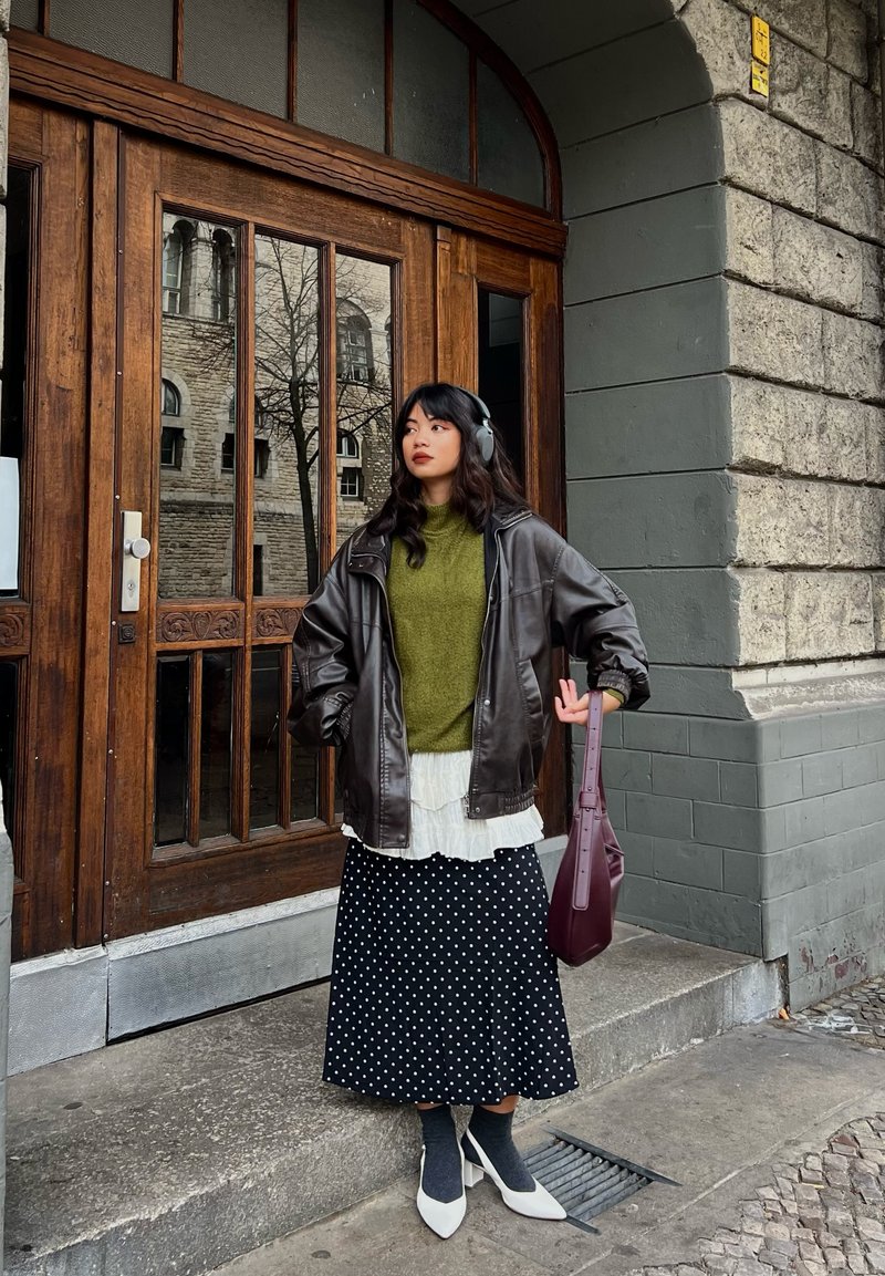 Giacca oversize in pelle marrone, maglione verde a maglia, camicetta bianca con volant, gonna nera a pois, calzini grigi, tacchi bianchi, borsa color borgogna scuro.