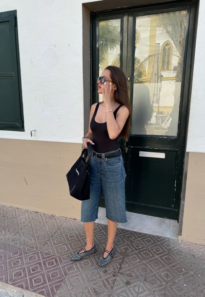 Mono negro, culottes de mezclilla, bolsa tote negra, zapatos planos con lunares. Cabello largo, gafas de sol, de pie junto a una puerta verde sobre un pavimento texturizado.