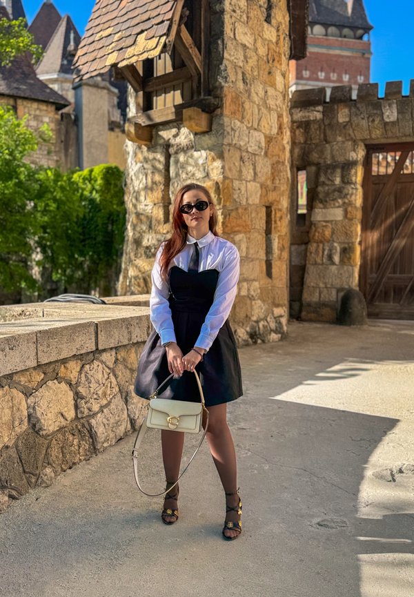 Mujer con vestido negro, camisa blanca y gafas de sol sosteniendo un bolso beige, de pie junto a una pared de piedra cerca de edificios de estilo castillo a la luz del sol.