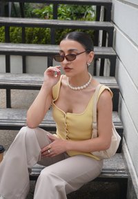 Žuti pleteni top s detaljima na dugmadima, bež široke hlače, biserna ogrlica i sunčane naočale, sjedi na crnim stepenicama na otvorenom.