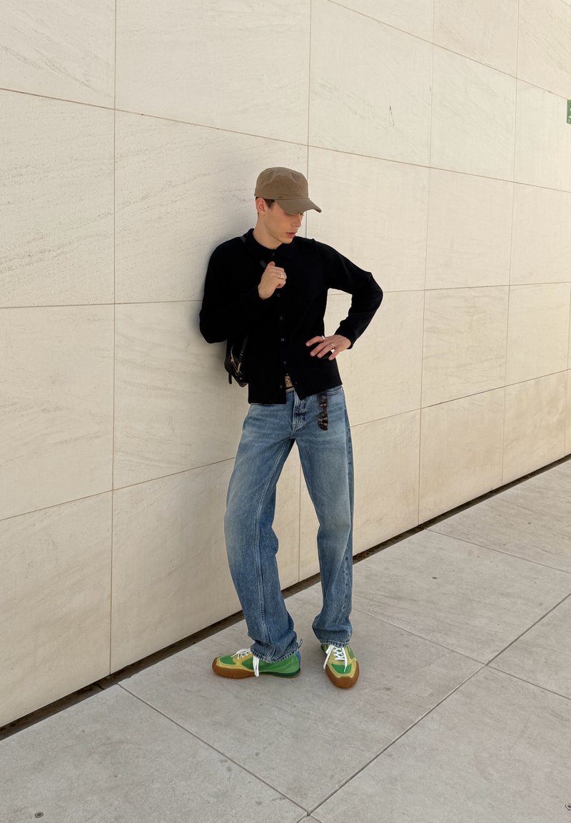Young man in a brown cap, black sweater, blue jeans, and green sneakers leaning against a beige tiled wall on a sidewalk.