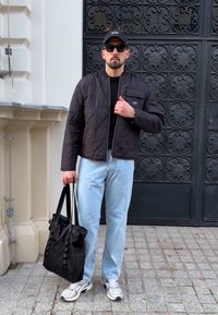 Černá prošívaná bunda, černý svetr, světle modré džíny, bílé tenisky, drží černou tašku přes rameno. Outfit doplňují sluneční brýle a kšiltovka.