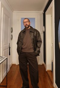 Brązowa skórzana kurtka na brązowym swetrze, w połączeniu z ciemnobrązowymi spodniami. Grube okulary i krótka broda. Drewniana podłoga i białe ściany.