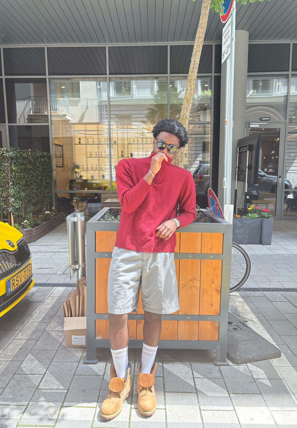 Pulôver de malha vermelho, calções cinzentos, botas castanhas com detalhes laranja, meias brancas; de pé ao lado de um vaso de madeira num ambiente urbano.