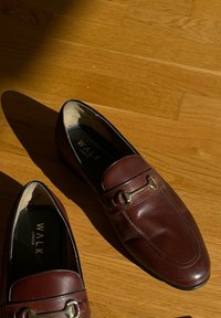 Pair of brown leather loafers with metal horsebit detail on wooden floor, lit by sunlight with visible shadows.