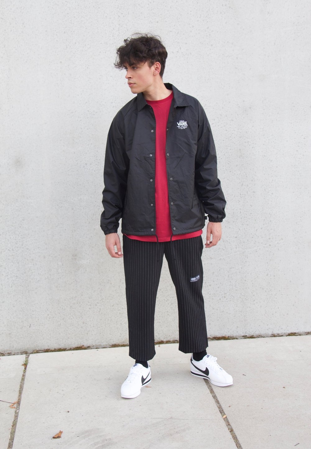 Black windbreaker with logo, red t-shirt, pinstriped pants, and white sneakers with black accents. Standing pose against a gray wall.