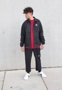 Black windbreaker with logo, red t-shirt, pinstriped pants, and white sneakers with black accents. Standing pose against a gray wall.