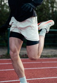 Juoksija kevyissä, vaalean kermavärisissä shortseissa mustan aluspaidan kanssa. Käyttää hanskoja ja harmaita sukkia, valkoiset ja oranssit urheilukengät.