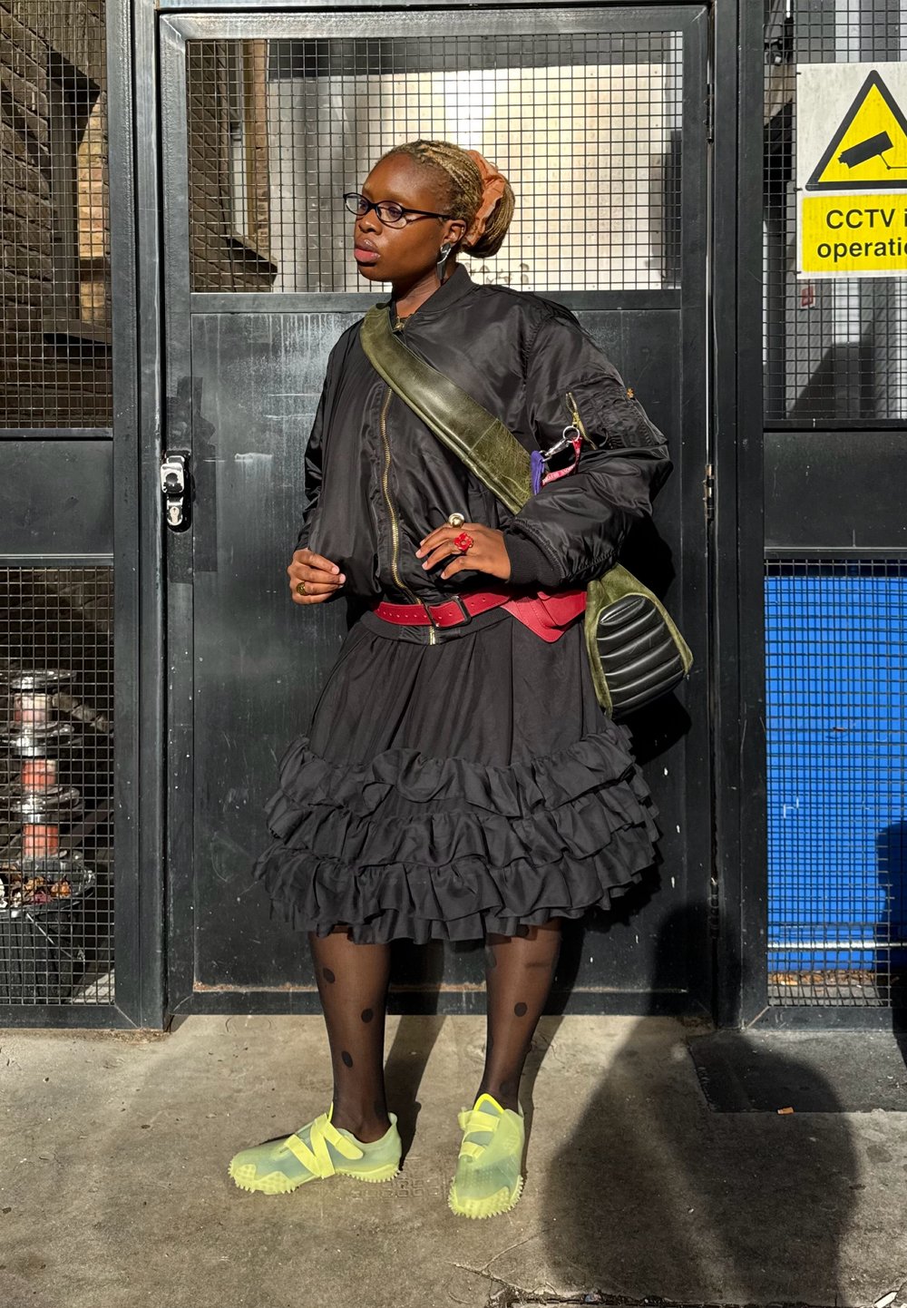 Čierna bomber bunda, červený opasok, vrstvená čierna sukňa s volánikmi, pančuchy s bodkami, žltá obuv a zelená kabelka cez rameno. Mestské prostredie.