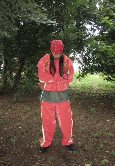 Chándal rojo y brillante con acentos naranjas; incluye una camiseta de malla verde debajo. Lleva zapatos negros y un pañuelo rojo. Fondo natural.