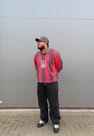 Hombre con camiseta de manga larga a rayas rojas y negras, pantalones cargo negros, gorra y gafas, de pie sobre suelo pavimentado frente a una pared gris.