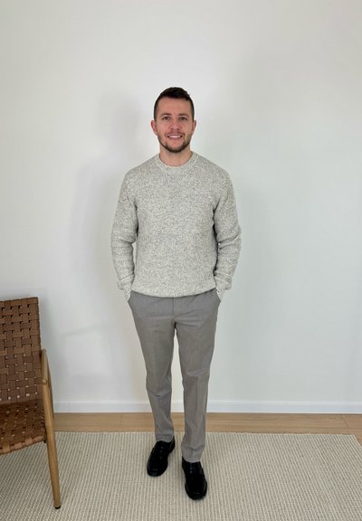 Hombre de pie en el interior, apoyado contra una pared blanca, vistiendo un suéter gris, pantalones grises y zapatos negros, con las manos en los bolsillos, junto a una silla de madera tejida.