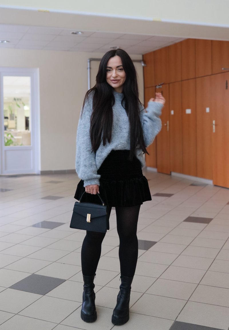 Pull gris duveteux ; jupe noire plissée en velours ; collants opaques noirs ; bottines noires à plateformes épaisses ; petit sac à main noir avec accent doré.