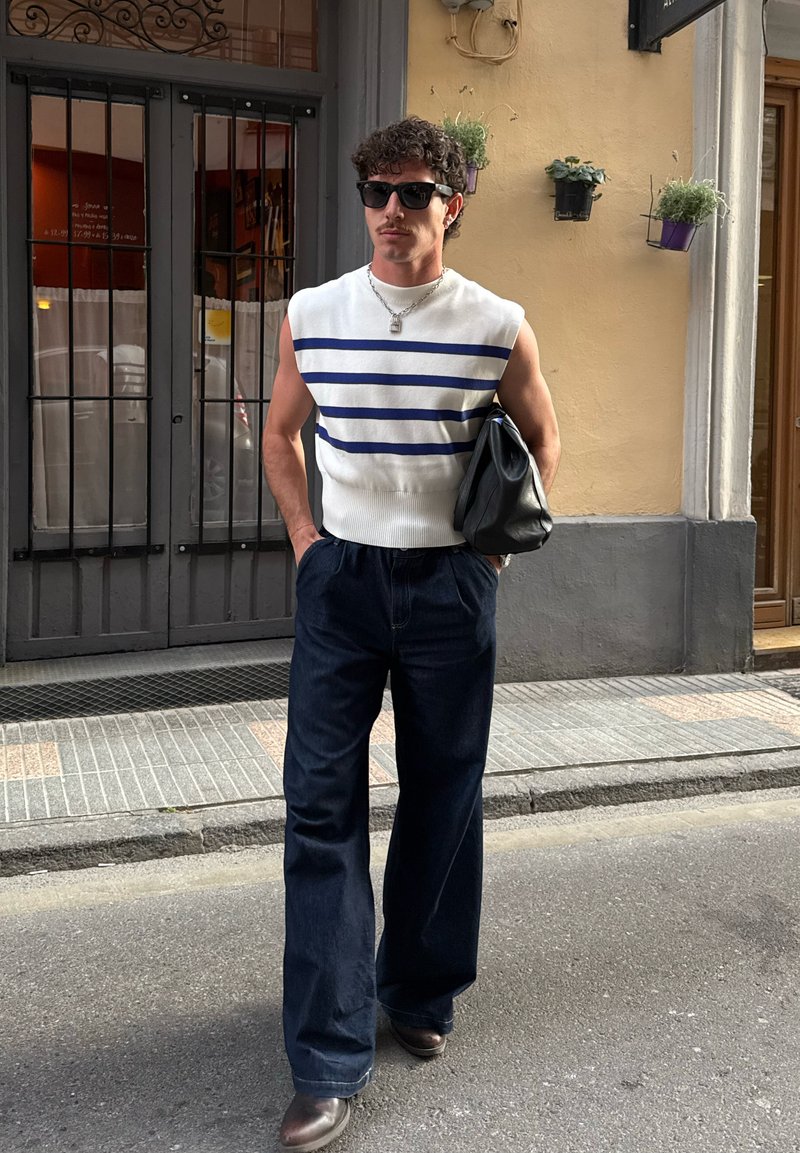 Homme portant des lunettes de soleil et un pull sans manches blanc avec des rayures bleues, un pantalon large foncé, tenant un sac noir dans une rue en ville.