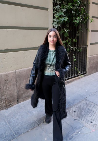 Abrigo de piel sintética negra con mangas largas, blusa gris a cuadros y pantalones negros. Modelo camina por una calle de piedra con plantas verdes de fondo.
