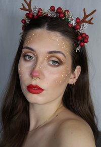 Porträtaufnahme einer Person mit langem braunem Haar, die ein Blumenhaarband mit roten Beeren und Geweihakzenten trägt. Helle rote Lippen und eine rosafarbene Nase.