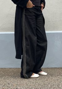 Pessoa vestindo calças largas pretas com riscas brancas nas laterais, casaco preto e sapatos brancos rasos, de pé em um chão texturizado ao lado de uma parede bicolor.