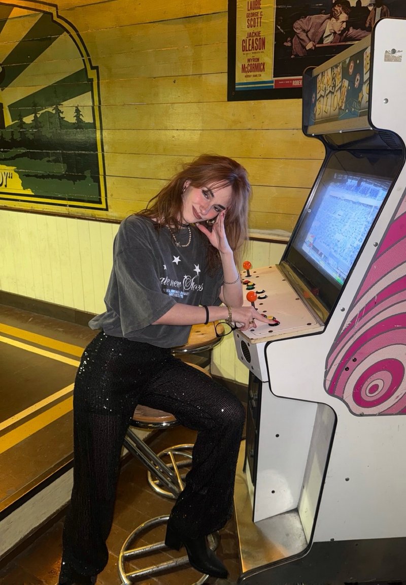 Cabinet de jeu d'arcade au style rétro avec un panneau coloré, équipé d'un joystick et de boutons. Pantalon large noir à sequins et chemise grise visibles.