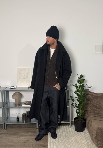 Hombre con gorro negro, abrigo largo negro, pantalones oscuros y zapatillas de deporte, de pie en una habitación minimalista con una planta, una estantería y un cartel de "Se busca".