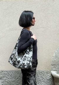 Čierna a sivá vzorovaná textilná taška na rameno s širokým dnom a dlhými rukoväťami, nesená na ramene na neutrálnej stenovej pozadí.