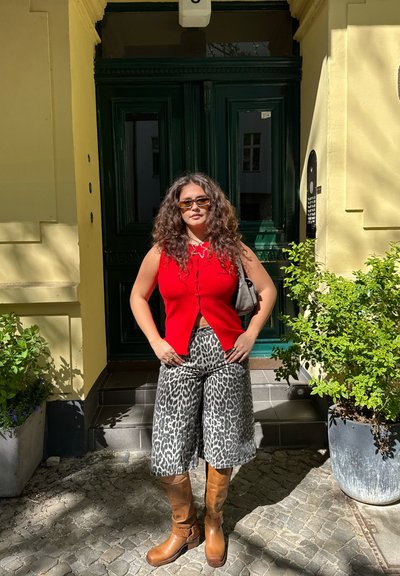 Cárdigan sin mangas rojo, culottes con estampado de leopardo, botas marrones hasta la rodilla. Puerta verde y pared amarilla al fondo. Plantas en macetas cerca.