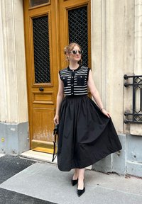 Abito nero con gonna ampia e top senza maniche a righe, dotato di bottoni e due tasche sul petto. Indossato con décolleté nere a punta e occhiali da sole.