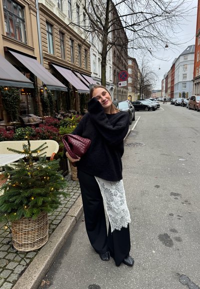Suéter negro extragrande, pantalones negros anchos con detalle de encaje blanco, bolso burdeos, entorno urbano al aire libre con vegetación y señales de tráfico.
