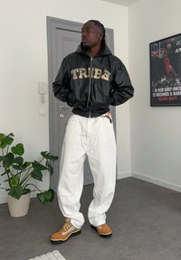 Veste aviateur en cuir noir avec le mot « TRIBE » en fauve, pantalon large blanc cassé, et bottes de randonnée fauve avec des lacets jaunes. Cadre intérieur avec plante.
