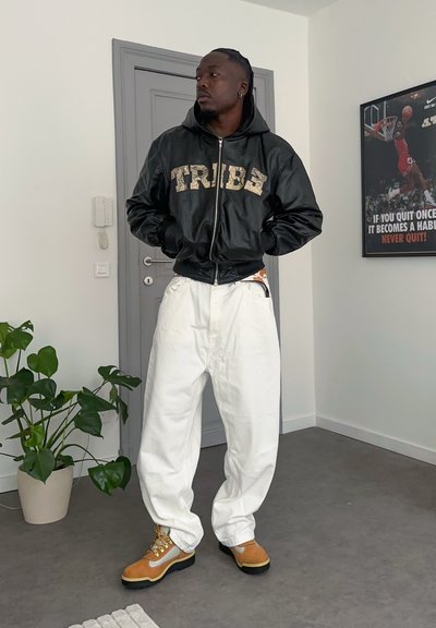 Chaqueta bomber de cuero negro con "TRIBE" en color marrón claro, pantalones blancos de gran tamaño y botas de senderismo marrón claro con cordones amarillos. Ambiente interior con planta.