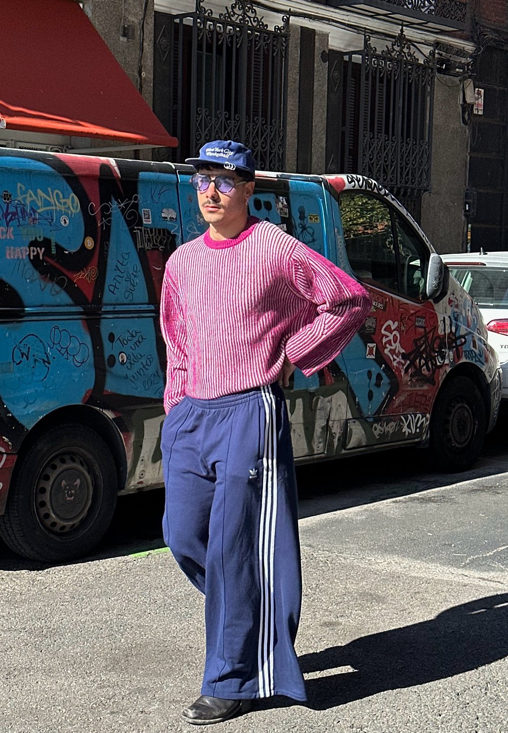 Uomo che indossa un maglione a righe rosa e pantaloni larghi blu con strisce bianche. Gli accessori includono occhiali da sole e un cappellino blu.
