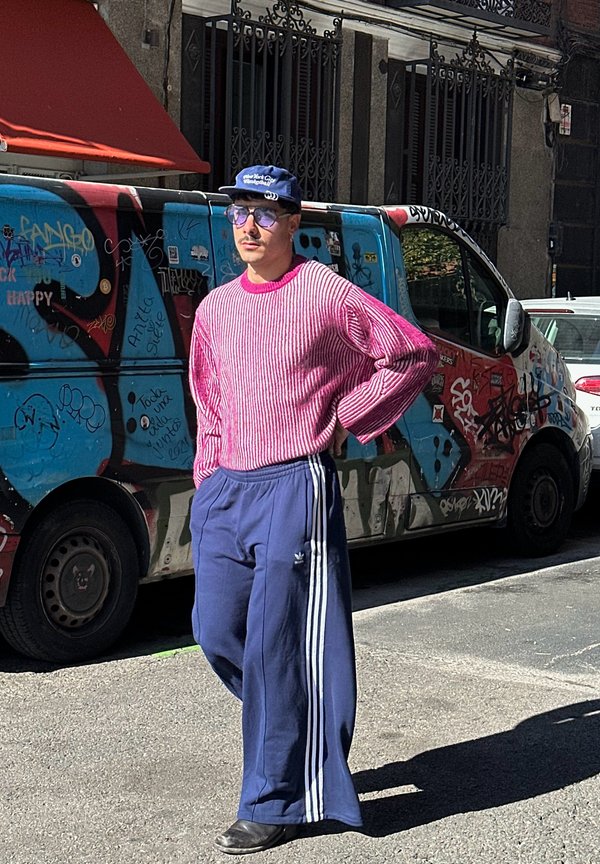 Man wearing a pink striped sweater and navy wide-leg pants with white stripes. Accessories include sunglasses and a blue cap.