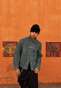 Jeansowa kurtka w wyblakłym niebieskim kolorze, z wysokim kołnierzem i guzikami z przodu. Czarna czapka beanie, czarne spodnie i dekoracyjny element na boku. Pomarańczowe tło.