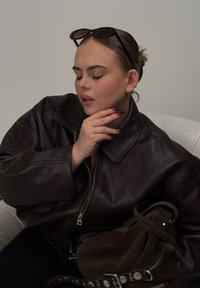 Geacă din piele maro cu guler mare și fermoar, asortată cu o geantă din suede închis. Unghii lungi, ombre și ochelari de soare așezați pe cap.