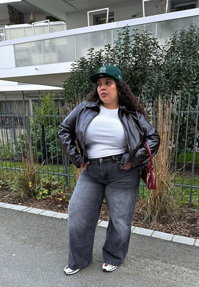 Chaqueta de cuero negra, camiseta blanca ajustada, vaqueros grises de cintura alta, gorra verde, bolso rojo y zapatillas blancas con acentos rojos y negros.