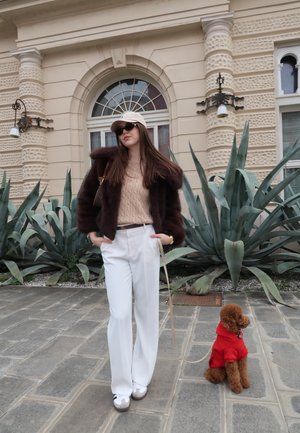 Mujer con gafas de sol, sombrero beige, chaqueta de piel marrón, pantalones blancos, de pie sobre un pavimento de piedra junto a un pequeño perro marrón con un suéter rojo.