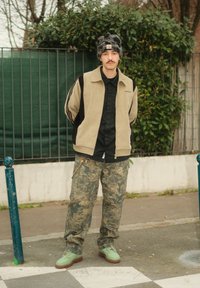 Veste en velours côtelé beige et noire, chemise noire, pantalon camouflage, chaussures vertes et bonnet à motifs. Debout devant un fond vert.