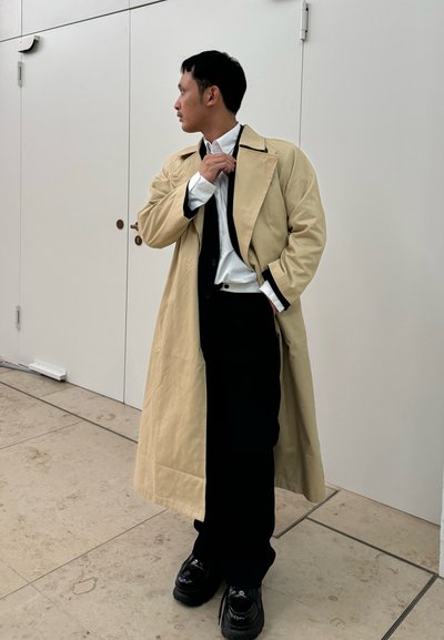 Trench coat beige con solapas anchas, sobre una camisa blanca y una corbata negra, combinado con pantalones negros y zapatos brillantes, de pie en un entorno minimalista.