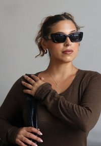 Mujer con cabello castaño recogido en un moño, usando gafas de sol oscuras, camiseta de manga larga marrón, sosteniendo una correa de cuero negra sobre su hombro.