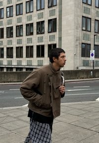 Jaunas vyras su trumpais tamsiais plaukais, dėvintis rudą švarką ir languotas kelnes, vaikšto miesto šaligatviu netoli daugiabučio pastato.