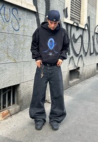 Melna kapuce ar zilu grafisku sejas dizainu, apvienota ar pārāk lielām melnām džinsu biksēm un melnām kurpēm, pret pelēku sienu ar grafiti.