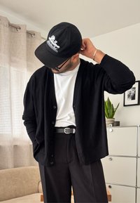 Mann trägt schwarze Mütze, schwarzen Cardigan, weißes Hemd und schwarze Hose, richtet die Mütze drinnen mit Vorhängen, Pflanze und gerahmtem Foto im Hintergrund.