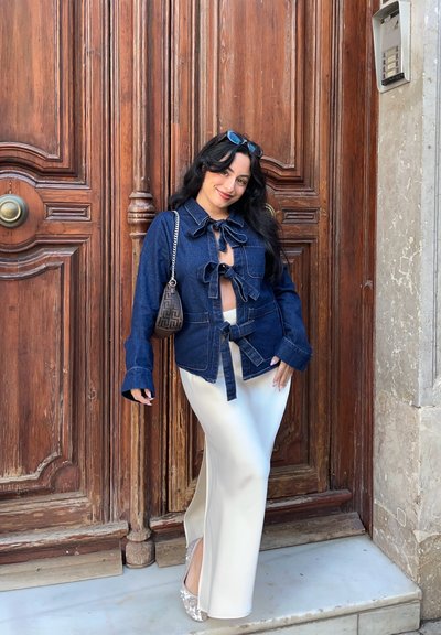 Chaqueta de mezclilla con lazo frontal, mangas largas y lavado en azul claro; combinada con una elegante falda de satén blanca con una abertura lateral. Tacones blancos adornados.