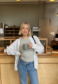 Ljusrandig skjorta över en beige topp, ljusblå vida jeans, hållande en grå kopp i en modern kafémiljö. Trädisk i bakgrunden.
