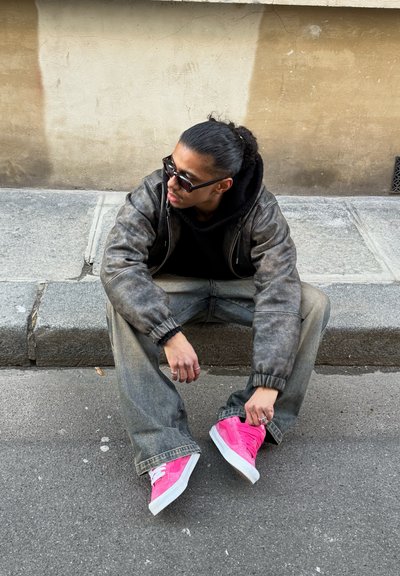 Joven con cabello oscuro en una coleta, lleva gafas de sol, chaqueta gris, vaqueros holgados y zapatillas rosas brillantes sentado en un bordillo de hormigón.