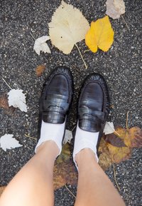 Crne kožne mokasine s teksturiranim dizajnom i šivenim detaljima, uparene s bijelim čarapicama do gležnja, na pozadini jesenskog lišća.