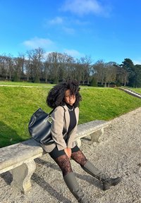 Sort tote bag med et rundt håndtag, grå blazer over sort top, mønstrede sorte strømpebukser med grå knæhøje støvler, siddende på en stenbænk.