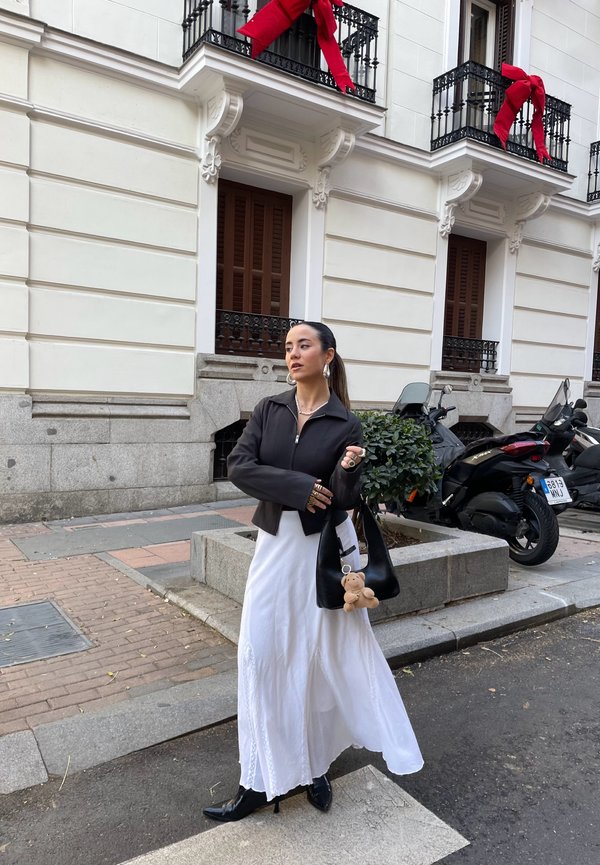 Chaqueta corta negra con cremallera, vestido maxi blanco con bordado, bolsa de mano negra con un charm de oso de peluche, y botas de tacón negras brillantes.