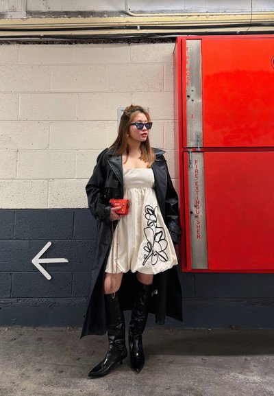 Mujer con vestido blanco de flores, abrigo de cuero negro y botas, de pie junto a una pared cerca de un gabinete rojo de manguera contra incendios y extintor con una señal de flecha hacia la izquierda.