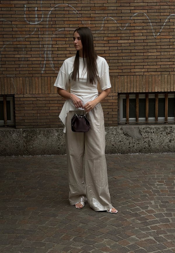 Mujer con cabello largo y oscuro está de pie en una calle empedrada, lleva una blusa blanca, pantalones sueltos beige, sandalias blancas y sostiene un bolso pequeño y oscuro.