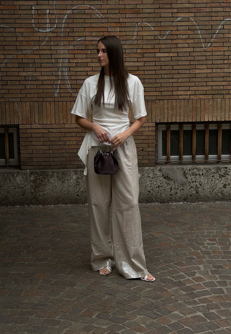 Femme aux longs cheveux foncés se tient dans une rue pavée portant un haut blanc, un pantalon ample beige, des sandales blanches, tenant un petit sac à main sombre.